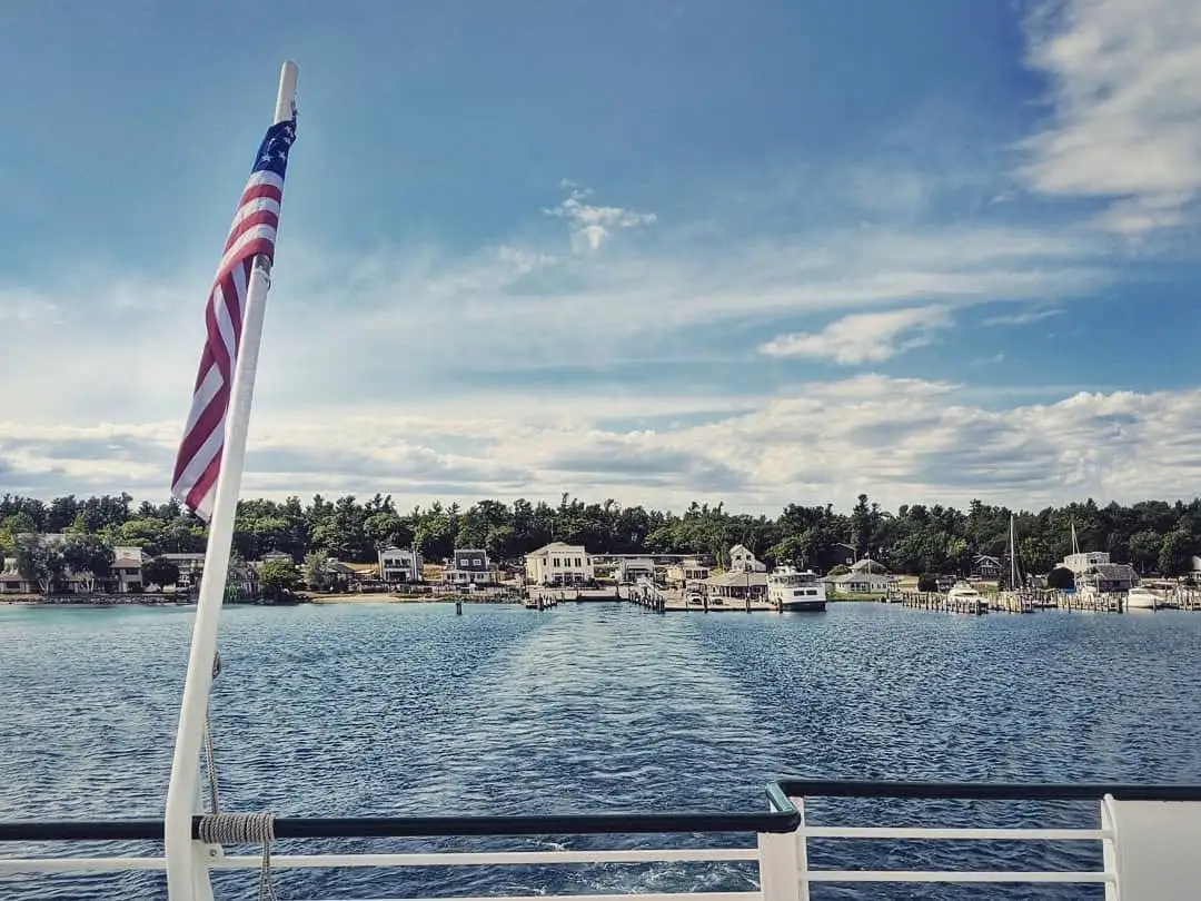 View from a boat across rippling water toward a tree-lined waterfront with docks, homes, and an American flag.