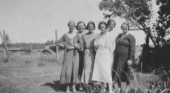 Six women pose together outdoors in a grassy field, with a fence, trees, and buildings behind them.