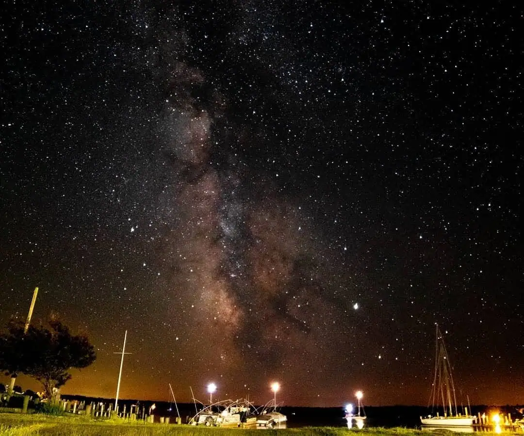The Milky Way rises above illuminated docks, boats, and a sailboat at night.