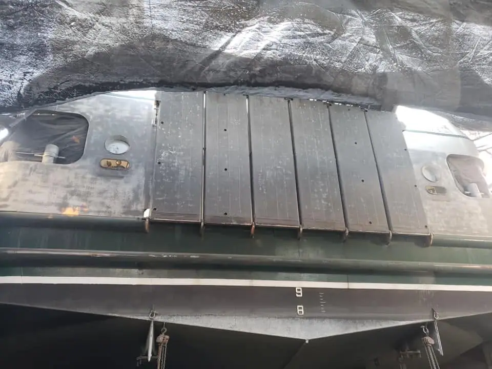 Close view of a weathered ferry hull with a large hinged panel, portholes, and hanging chains.