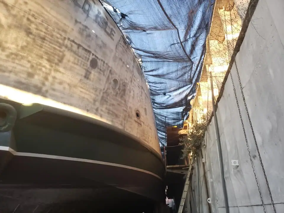 A large vessel hull is being maintained beneath a blue tarp in an enclosed work area.