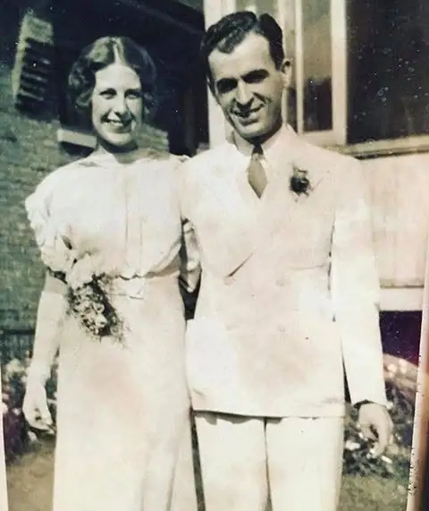 A smiling bride and groom stand outdoors in formal wedding clothing beside a building.