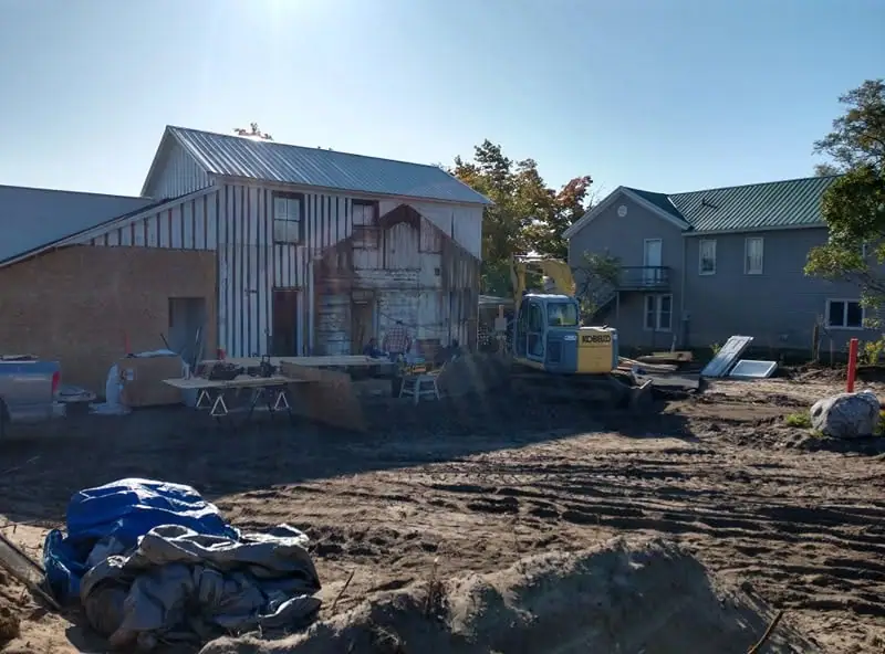 Construction work in front of a weathered barn and nearby house, with a compact excavator and building materials visible.