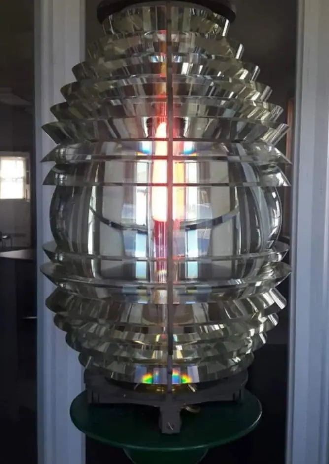 A large tiered Fresnel lens with ribbed glass panels and a central illuminated lamp stands in an indoor display.