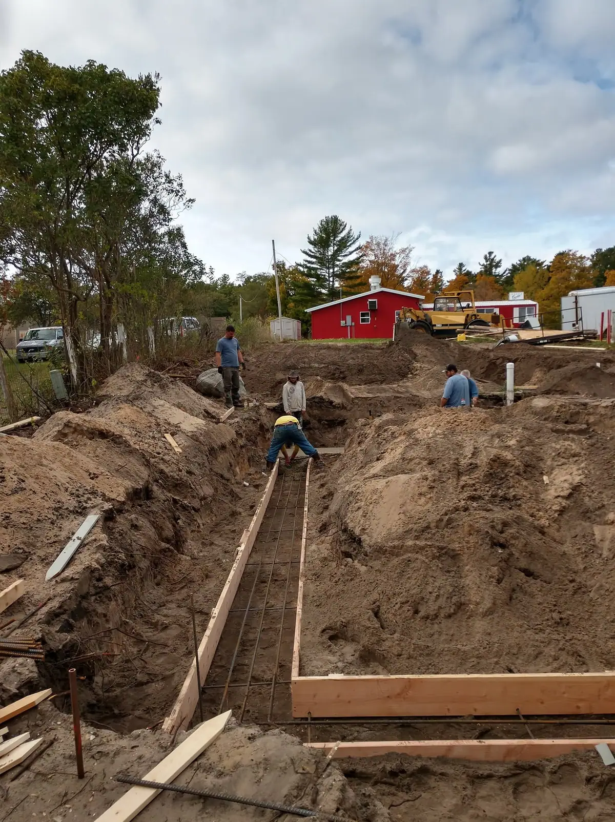 Workers prepare rebar and wooden forms in an excavated construction trench beside a tracked loader.