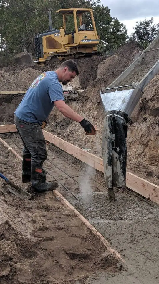 A worker pours concrete into a rebar-reinforced wooden form beside a tracked bulldozer.