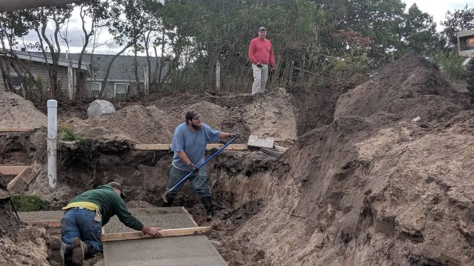 Three workers build a concrete structure in a deep excavation beside a house.