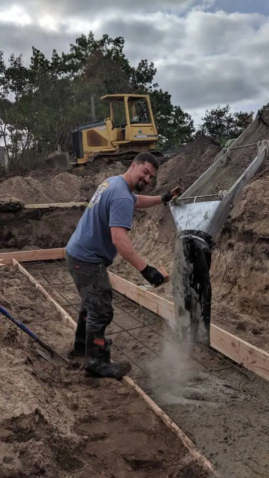 A worker pours wet concrete into a rebar-reinforced wooden form while a bulldozer stands behind him.