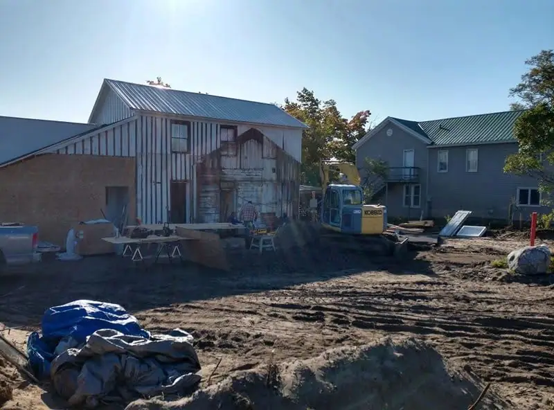 A barn-like building under renovation beside an excavator and a gray house.