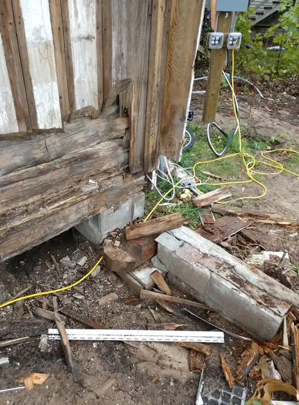 Close view of a severely decayed wooden structure with exposed wiring, concrete supports, and scattered debris.