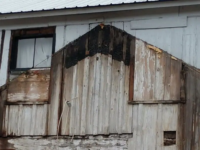 Close view of a weathered wooden structure with peeling paint, boarded openings, and a small window.