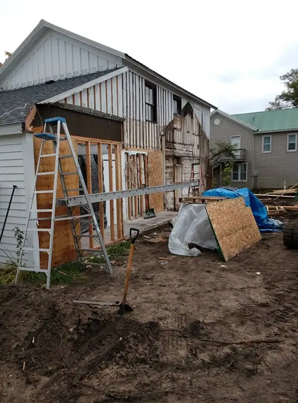 House with exposed framing, ladder, construction materials, and a shovel in a muddy yard.