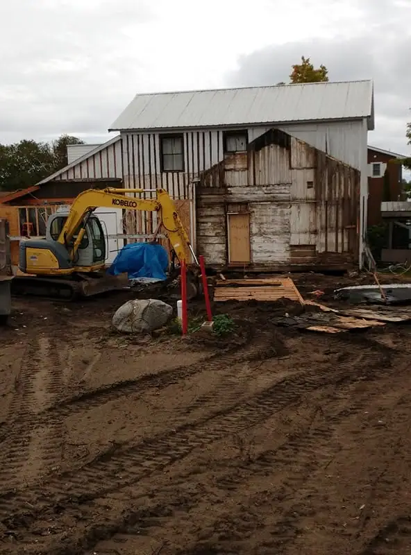 An excavator stands beside a weathered wood-sided building amid muddy ground and scattered demolition materials.