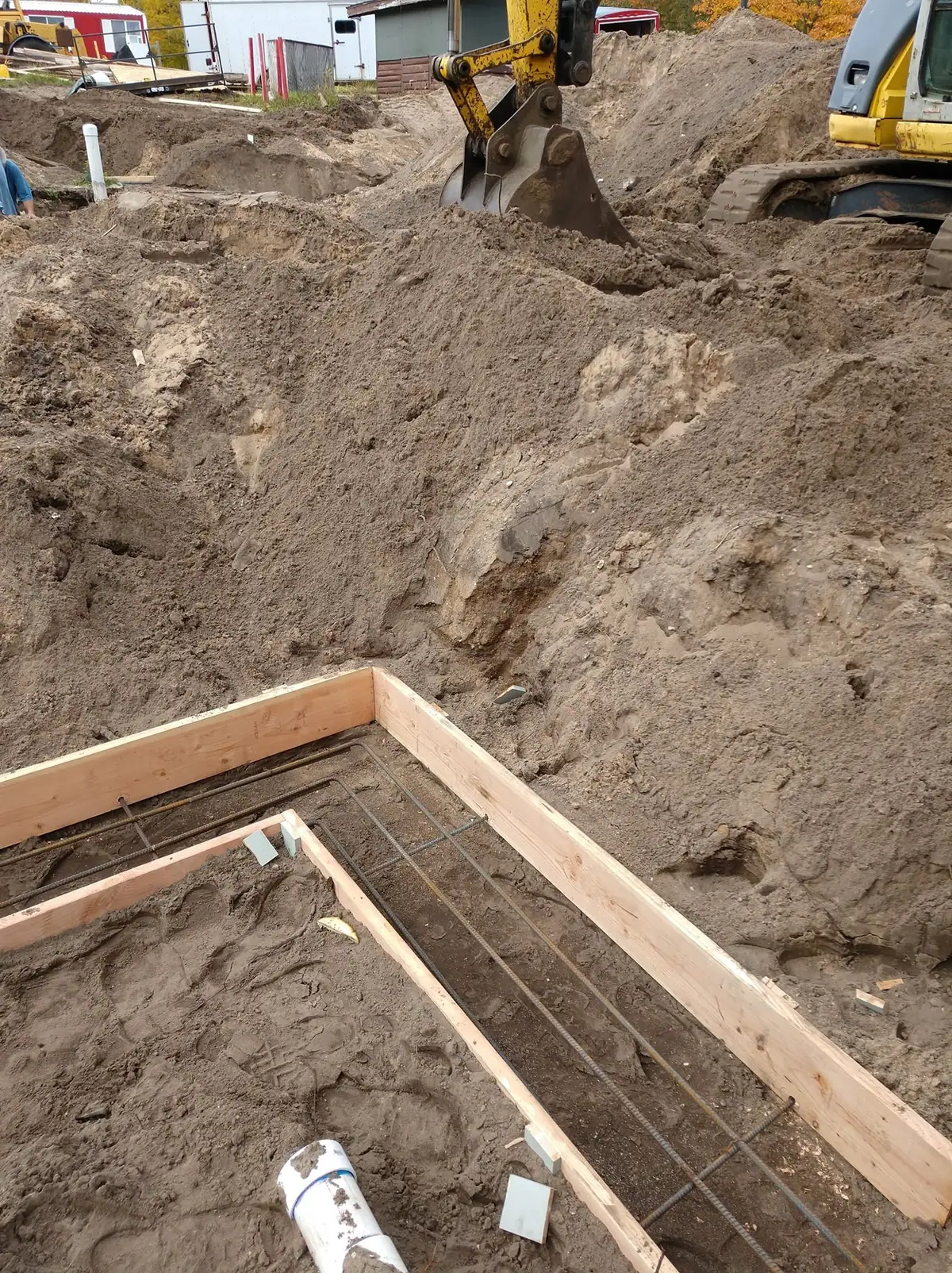 Excavator works beside wooden foundation forms filled with rebar at a construction site.