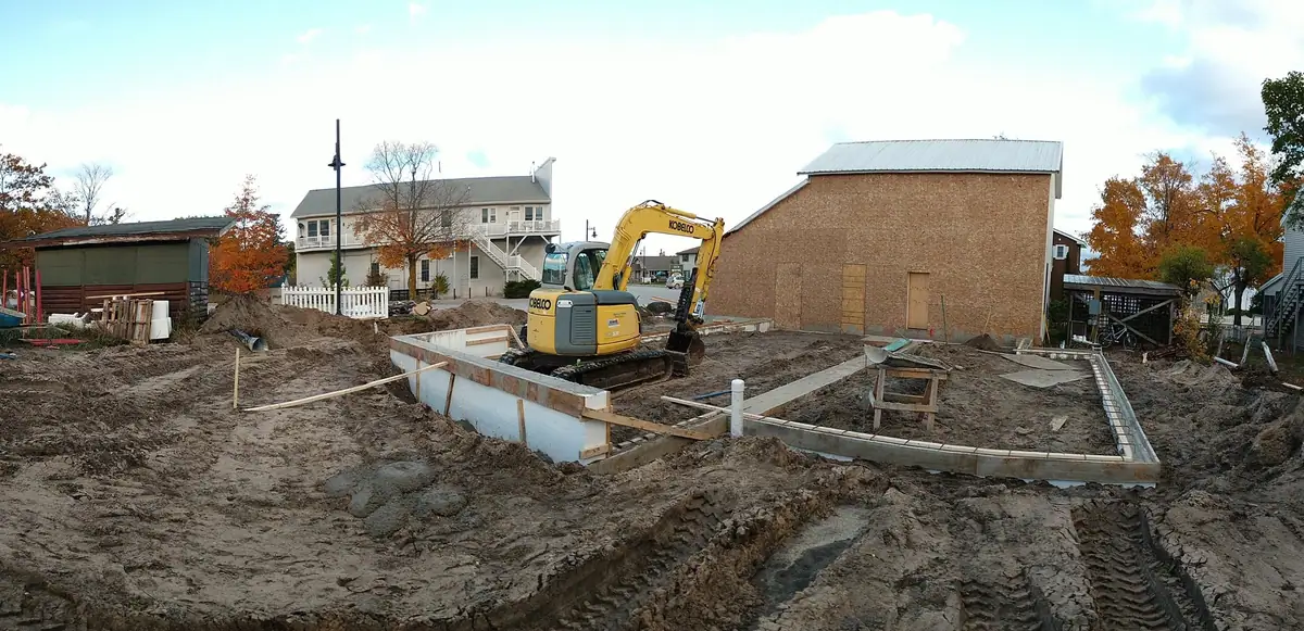 An excavator sits within a partially built foundation amid muddy ground and surrounding buildings.