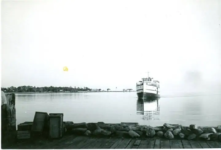 Black-and-white photograph of a ferry approaching a wooden dock on calm water.