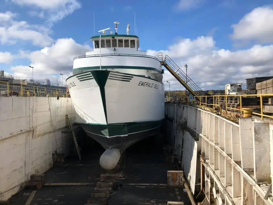 A white-and-green ferry rests bow-first in a concrete dry dock beneath a blue, cloud-filled sky.