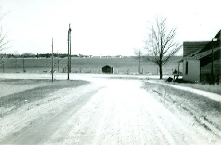 A snow-covered road leads toward a small outbuilding, open fields, utility poles, and a building at right.