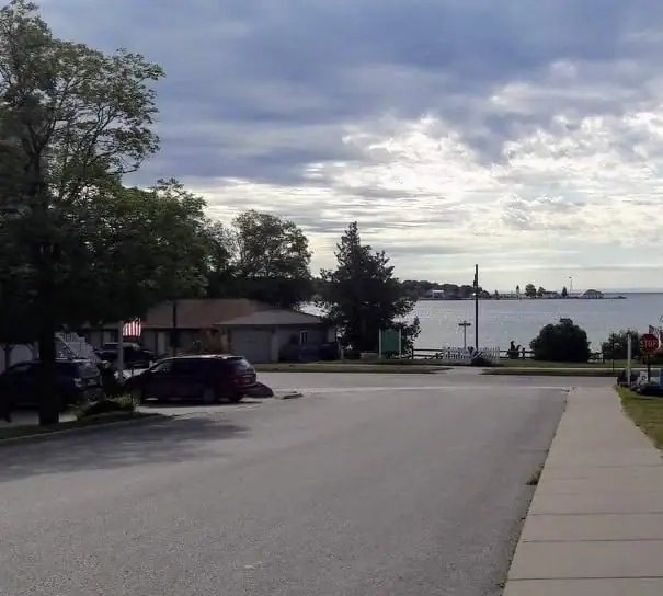A paved street leads toward a waterfront past a house, parked cars, trees, and a stop sign.