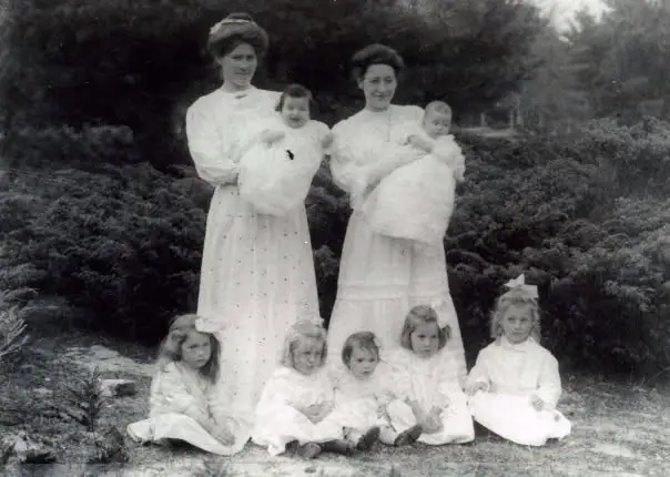 Two women holding babies stand behind five seated children in an outdoor group portrait.