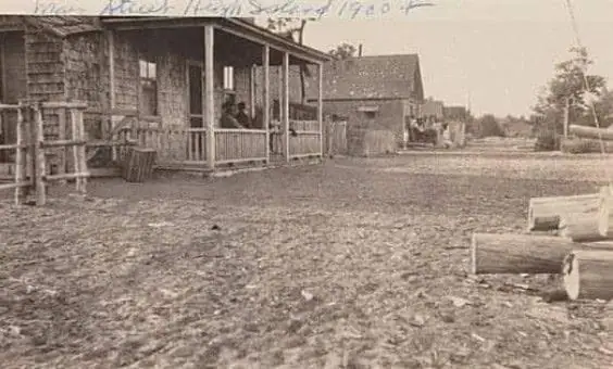 Sepia photograph of a wooden house and porch beside an unpaved street, with stacked logs in the foreground.