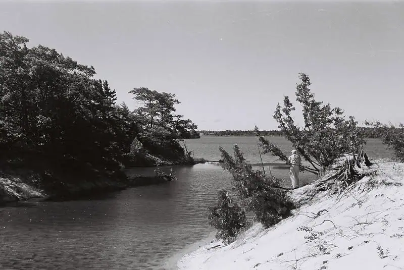 Black-and-white photograph of a person fishing beside a wooded water channel and sandy bank.