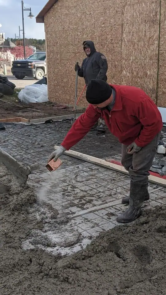 Two workers pour and spread concrete over a rebar-reinforced slab beside a sheathed building.