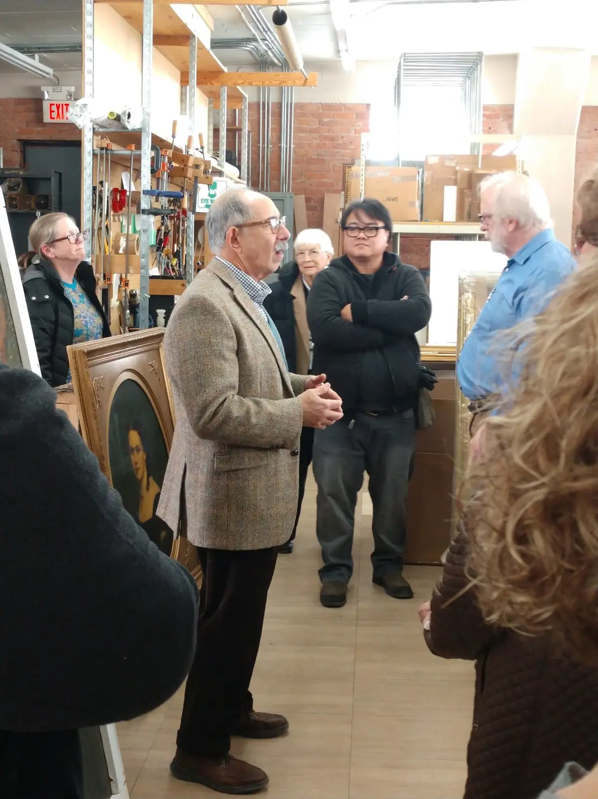 A man speaks to a small group in a workshop containing tools, shelving, boxes, and framed artwork.