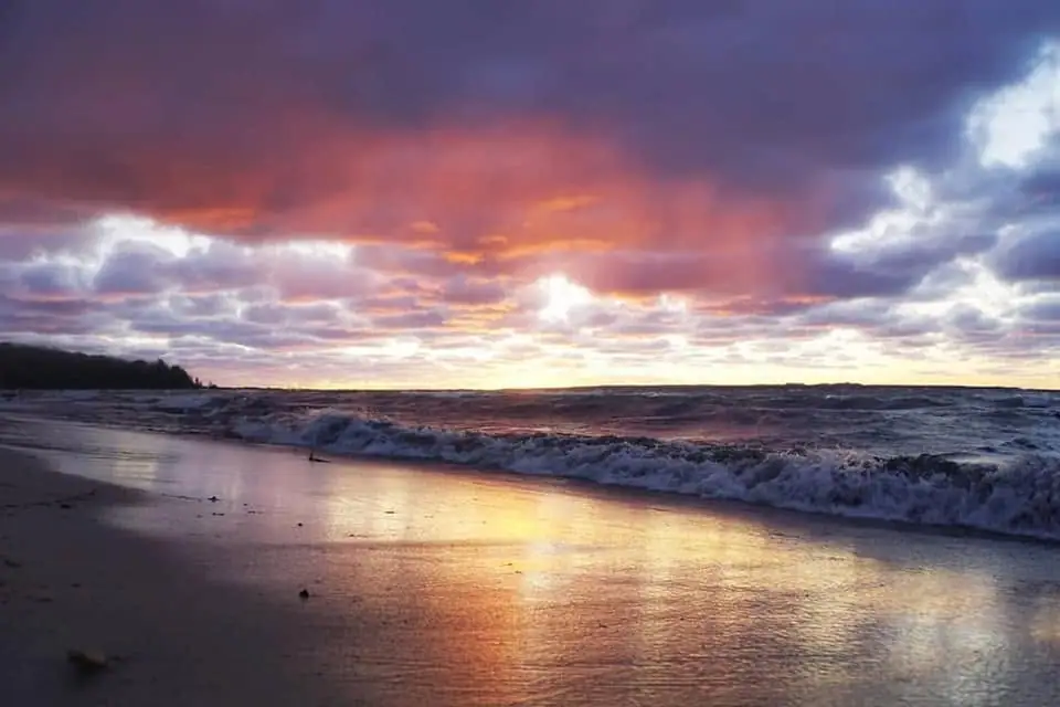 Sunset light reflects across wet sand as waves roll onto a beach beneath layered clouds.