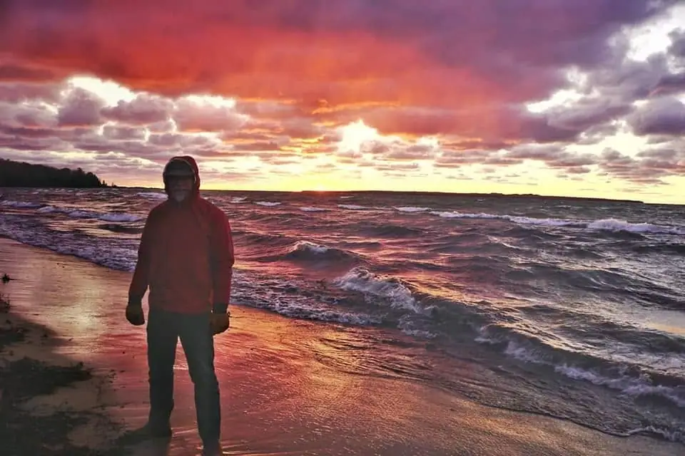 A hooded person stands on wet sand beside breaking waves beneath a vivid sunset sky.