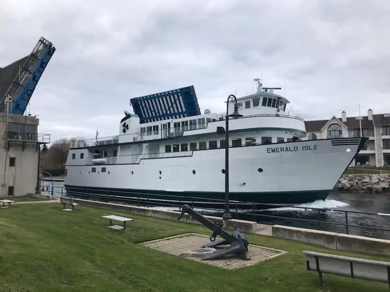 A white ferry passes a waterfront quay, with raised loading ramps and an anchor sculpture in the foreground.