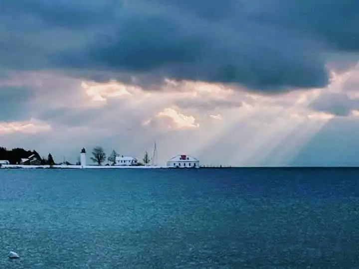 Lighthouse and shoreline buildings beside dark water beneath storm clouds and shafts of sunlight.