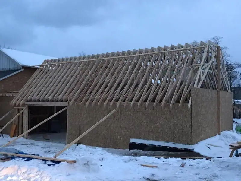 Partially constructed building with exposed wooden roof trusses and OSB walls in a snow-covered setting.