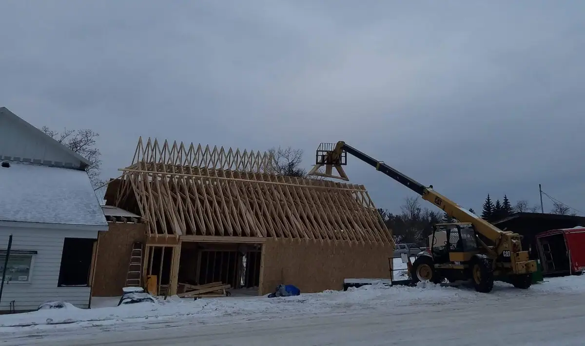 A yellow boom lift holds roof trusses above a snow-covered building under construction.