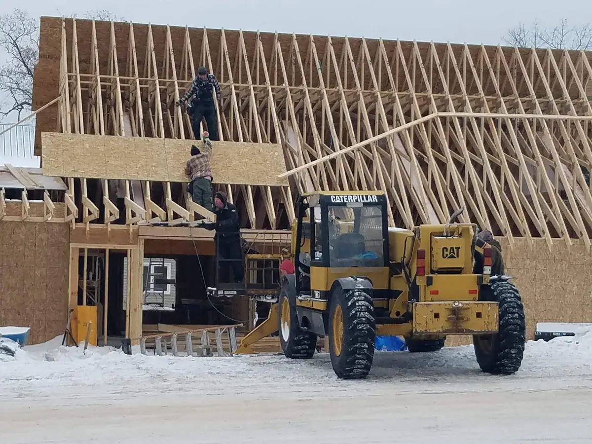 Workers install wood sheathing on a framed building beside a yellow loader in the snow.