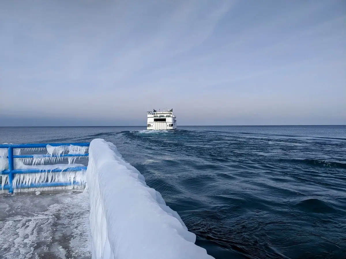 A ferry travels across open water while ice coats the foreground deck railing and hangs in icicles.