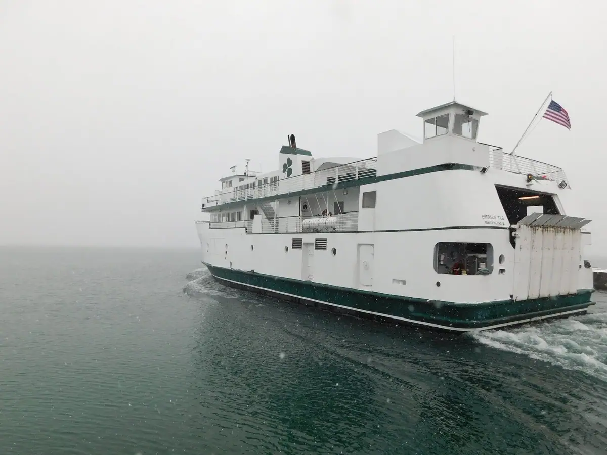 A white-and-green ferry moves through cold, hazy water with an American flag flying from its upper deck.