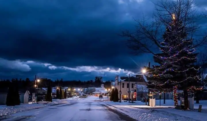 Snow-covered town street at night with illuminated buildings and a large decorated evergreen tree.
