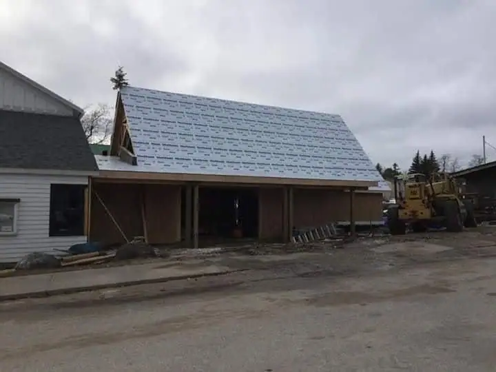 Partially constructed wood building with roof sheathing, a small evergreen, and a yellow excavator.