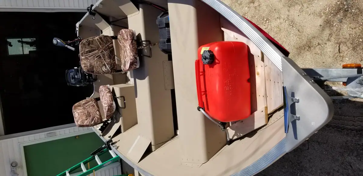 Small aluminum fishing boat on a trailer, with camouflage seats and a red fuel tank outside a garage.