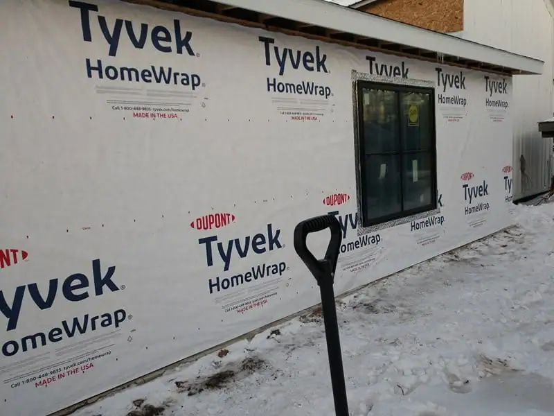 An exterior wall wrapped in Tyvek HomeWrap stands beside snow, with a window and shovel in view.