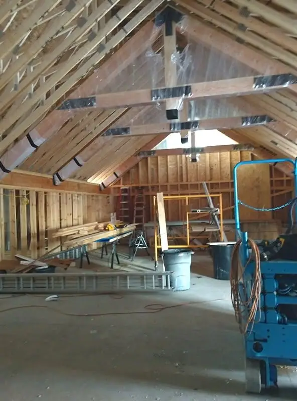 Interior of a timber-framed building under construction, with roof trusses, ladders, tools, and equipment.