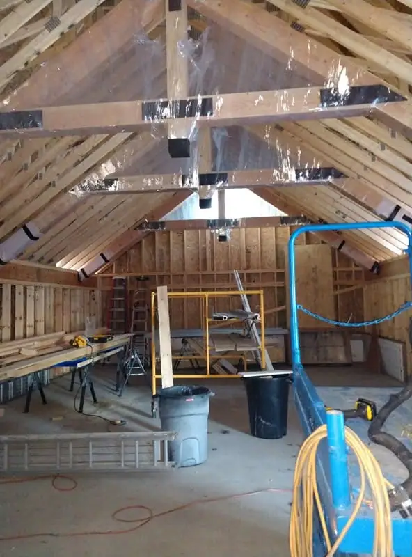 Unfinished timber-framed interior with roof rafters, plastic sheeting, ladders, tools, and construction materials.