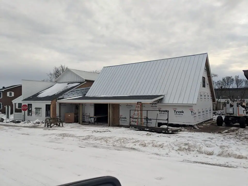 Snow-covered building under construction with a new metal roof, exposed wrap, ladder, and construction equipment.
