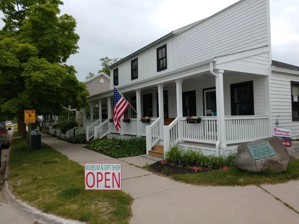 White two-story museum building with a porch, American flag, open sign, and roadside signs.
