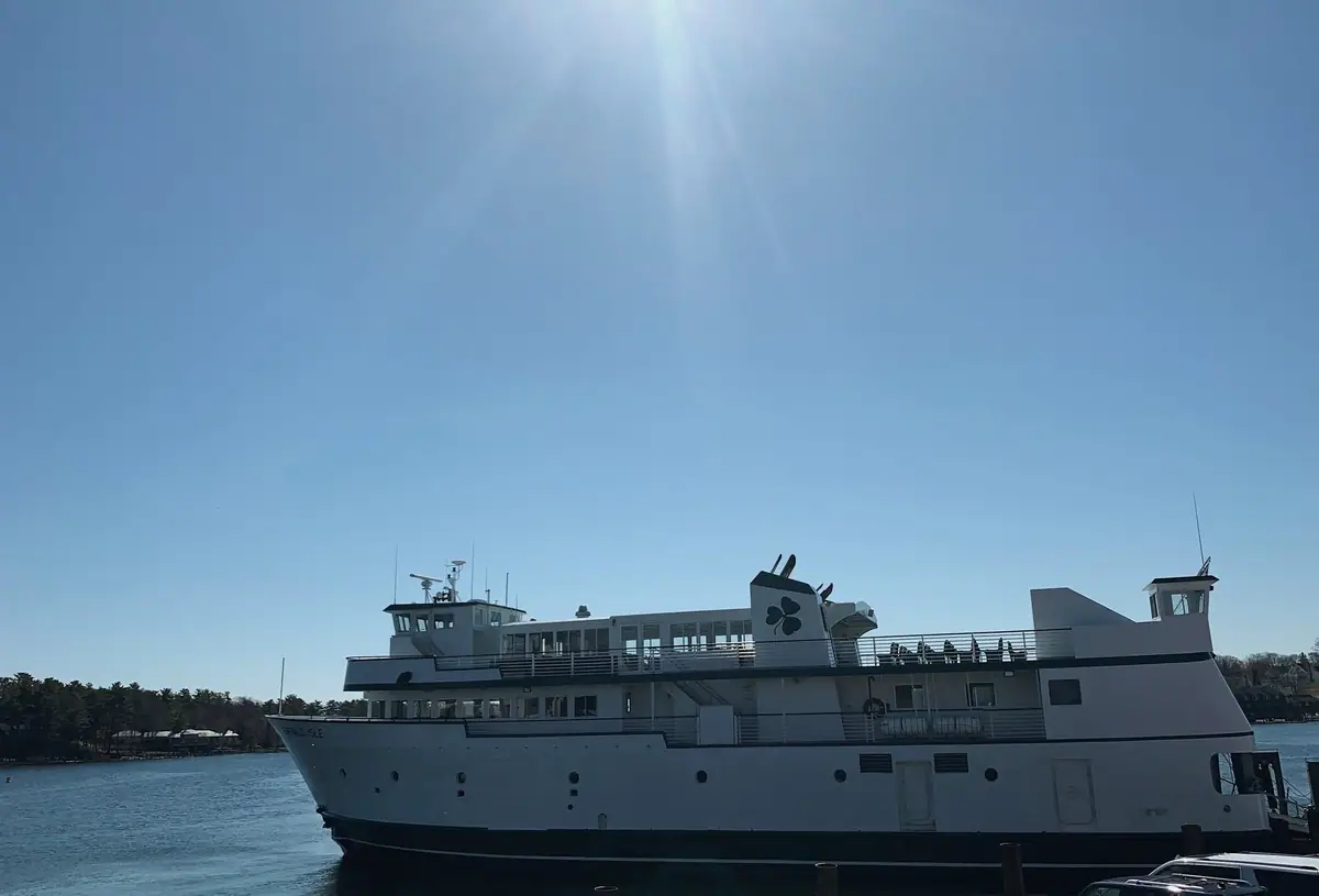 A white multilevel ferry with a shamrock emblem is beside a pier under a bright blue sky.