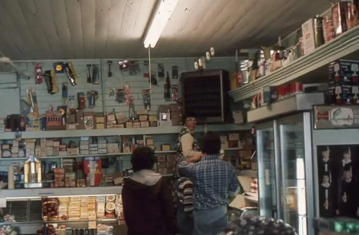 Three people stand at the counter of a small, densely stocked store interior.
