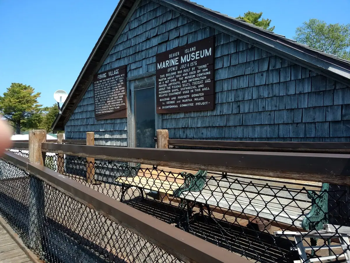 Weathered shingled museum building with two large informational signs, seen beyond a fenced wooden deck.