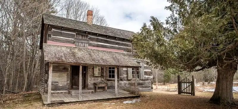 Weathered two-story log building with a porch, chimney, trees, and a wooden gate.