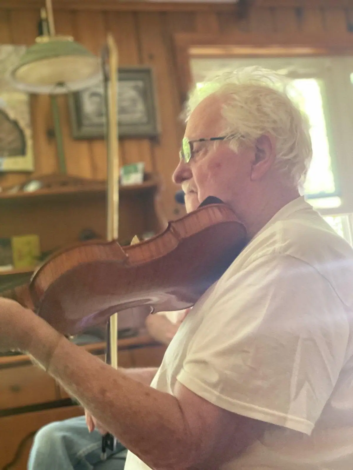 Older man in glasses playing a violin inside a wood-paneled room.